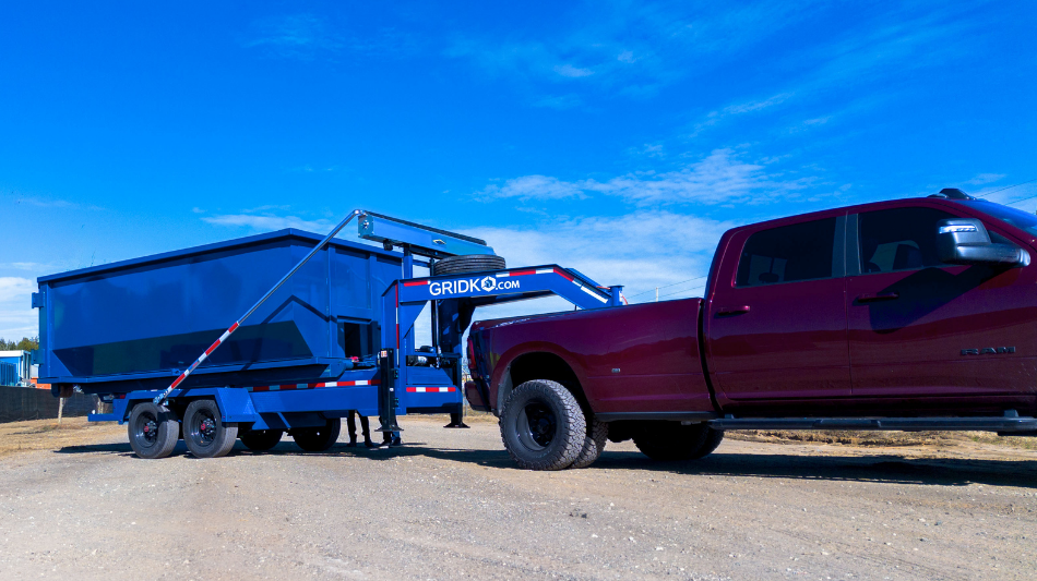 Blue roll-off dumpster container on a black Gridko gooseneck trailer, hitched to a maroon dually pickup truck on a dirt lot. 